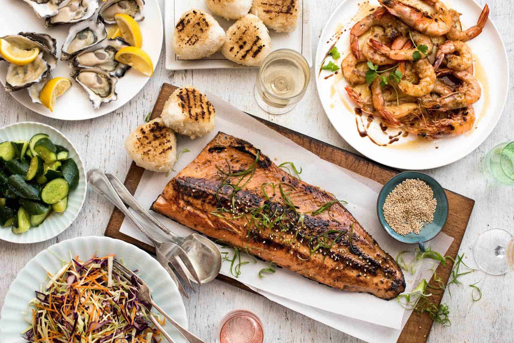 Overhead photo of an amazing Japanese BBQ menu with dishes including a BBQ Miso Salmon Side, grilled Asian prawns, grilled rice cakes, fresh oysters, Japanese cucumber salad and Asian Slaw.