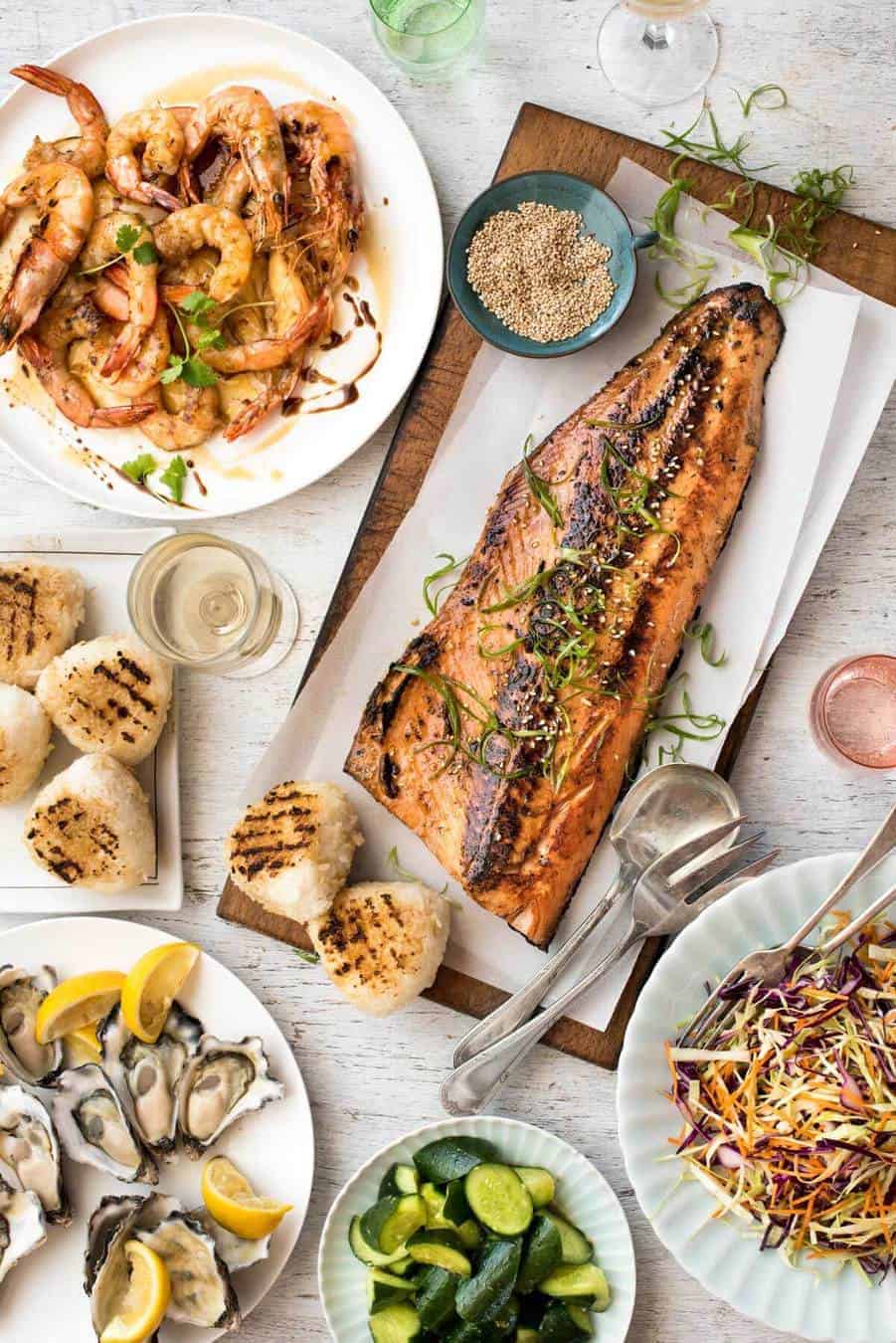 Overhead photo of an amazing Japanese BBQ menu with dishes including a BBQ Miso Salmon Side, grilled Asian prawns, grilled rice cakes, fresh oysters, Japanese cucumber salad and Asian Slaw.